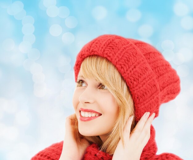 felicidade, férias de inverno, natal e conceito de pessoas - jovem sorridente de chapéu vermelho e cachecol sobre fundo de luzes azuis