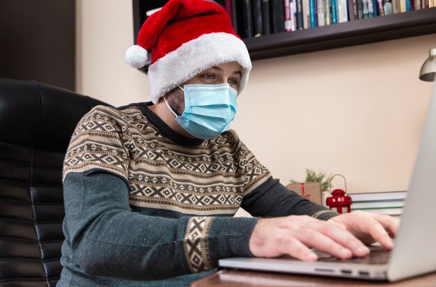 Felicitación de Navidad en línea. Hombre joven con sombrero de santa claus y mascarilla habla usando laptop para videollamadas amigos y niños. Navidad durante el coronavirus.