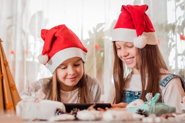 Felicitaciones en cuarentena. Las niñas con adornos navideños se ríen y se comunican con sus familias a través de una tableta.