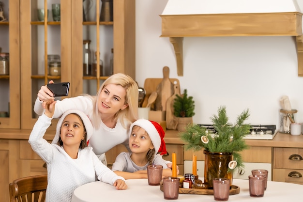 Felicitaciones familiares de Navidad en línea. Sonriente madre e hija rubia europea haciendo selfie foto en casa de disfraces de Navidad