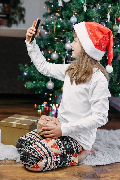 Felicitaciones de Navidad y Año Nuevo en línea. Linda chica sonriente usa el teléfono móvil para video chat en línea, llamadas a padres, familiares o amigos.