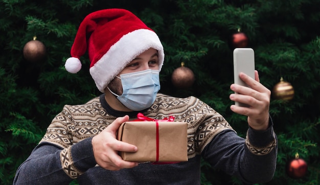 Felicitaciones de Navidad en línea. Retrato de hombre vestido con gorro de Papá Noel y máscara médica, dando caja de regalo con cinta roja, árbol de Navidad bokeh de fondo