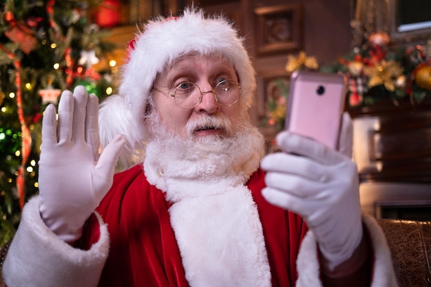 Foto felicitaciones navideñas en línea de santa papá noel sorprendido usando el teléfono móvil a distancia llamadas navideñas a personas y niños sentados cerca del árbol de navidad en casa feliz año nuevo covid 2021