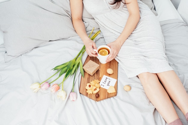 Felicitaciones a tu amada mujer en el día de San Valentín. Desayuno en la cama para una niña en camisón. Flores y regalo para el 14 de febrero.