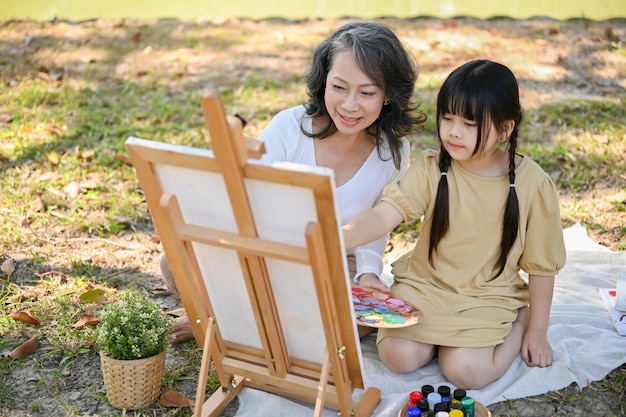 Feliz abuela asiática enseñando a su nieta a pintar acuarelas sobre lienzo