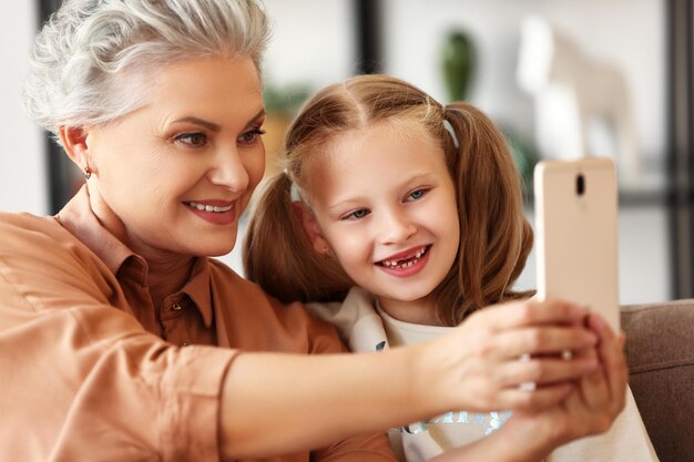 Feliz abuela familiar sonriendo y tomándose selfie con una alegre nieta mientras se sienta en un sofá en una acogedora sala de estar en casa