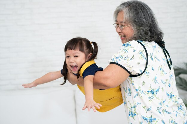 Feliz abuela mayor asiática sosteniendo y jugando con su nieta en la sala de estar