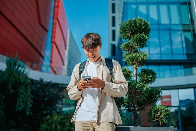 Feliz adolescente asiático participa de um delicioso bate-papo on-line em seu smartphone com mochila escolar por perto