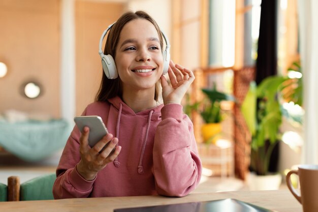 Feliz adolescente caucasiana usando fones de ouvido sem fio e usando o celular sentado na mesa em casa e sorrindo