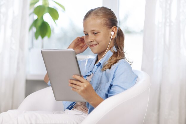 Feliz adolescente con ojos azules en una habitación luminosa, se sienta con una tableta y se comunica mediante una videollamada.