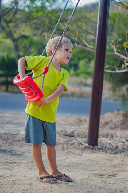 Feliz adorável menino loiro vai andar em um balanço do lado de fora no playground no parque Atividade de creche no horário de verão simples alegrias da infância