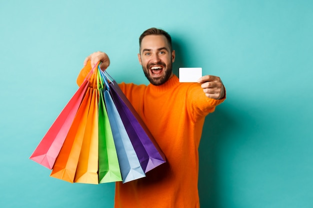 Feliz aduly hombre mostrando tarjetas de crédito y bolsas de compras, de pie contra el fondo turquesa