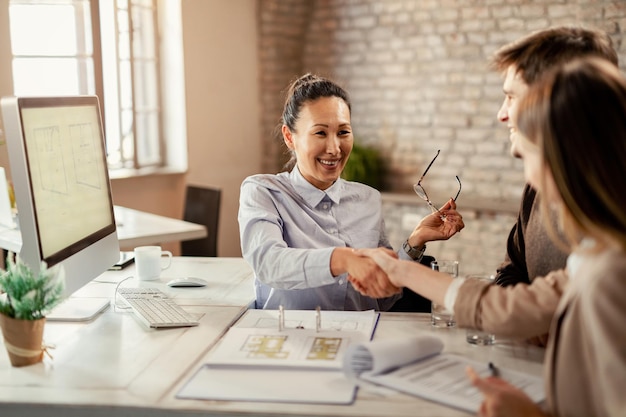 Feliz agente inmobiliario asiático estrechando la mano de una pareja durante una reunión en la oficina