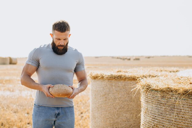 Feliz agricultor barbudo em jeans segurando pão fresco perfumado e de pé no campo ensolarado ao redor de fardos