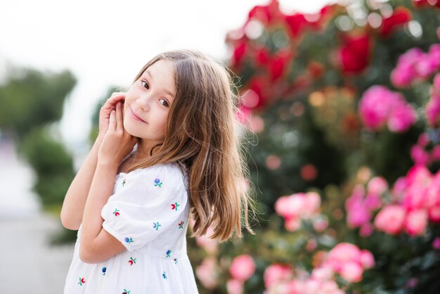 Feliz, alegre y adorable niña pequeña parada en el jardín del parque sobre arbustos florecientes de flor de rosa