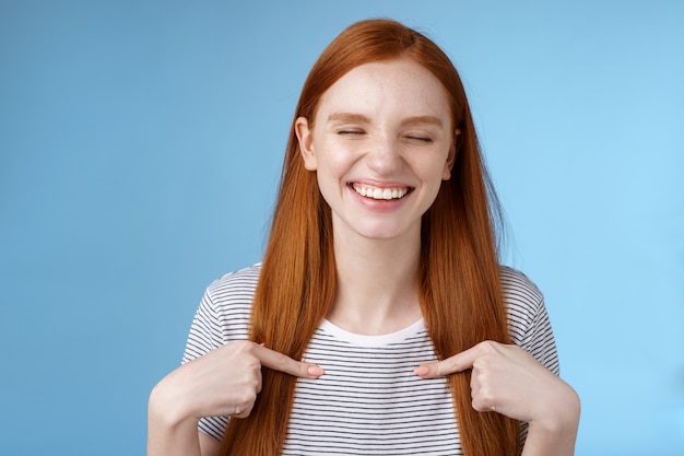 Feliz alegre alegre joven deportista pelirroja nominada sonriendo sorprendida riendo con alegría cerrar los ojos apuntando a sí misma elegida ganó el primer premio, de pie con fondo azul.