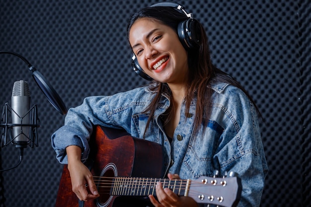 Feliz, alegre e sorridente do retrato de uma jovem vocalista asiática usando fones de ouvido com uma guitarra gravando uma música na frente do microfone em um estúdio profissional