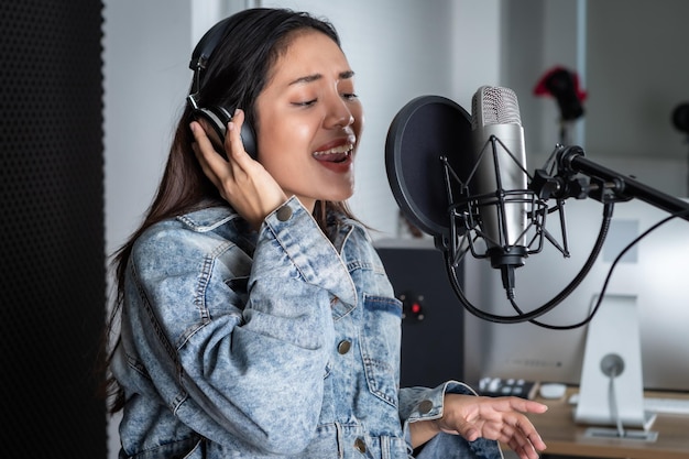 Feliz, alegre e sorridente do retrato do jovem vocalista asiático usando fones de ouvido, gravando uma música na frente do microfone em um estúdio profissional