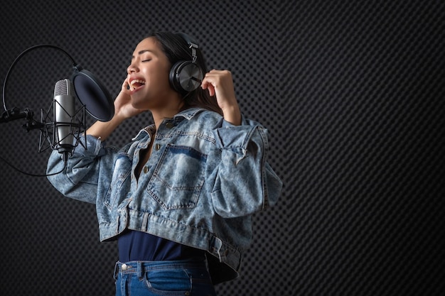 Foto feliz, alegre e sorridente do retrato do jovem vocalista asiático usando fones de ouvido, gravando uma música na frente do microfone em um estúdio profissional
