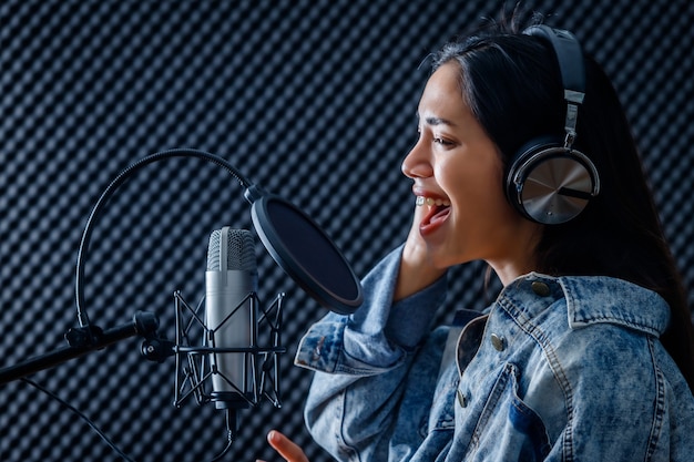 Feliz alegre e sorridente do retrato do vocalista jovem mulher asiática usando fones de ouvido, gravando uma música na frente do microfone em um estúdio profissional