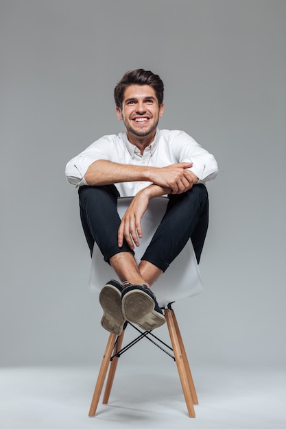 Feliz alegre homem bonito sentado na cadeira isolada em uma parede cinza