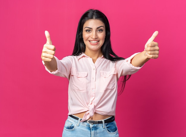 Feliz y alegre joven bella mujer en ropa casual mirando al frente sonriendo ampliamente mostrando los pulgares para arriba de pie sobre la pared rosa