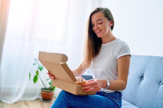 Foto feliz alegre joven satisfecha cliente mujer adicta a las compras sentado en el sofá en casa y abriendo la caja de paquete de cartón recibido, concepto de entrega de comercio de servicio fácil y rápido