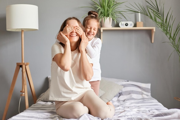 Feliz alegre madre alegre y su hijo divirtiéndose en el dormitorio niño abrazando a mamá cubriendo los ojos con las palmas jugando al escondite y enfermo en el dormitorio en casa