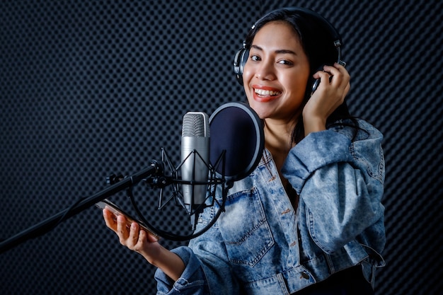 Feliz alegre muy sonriente del retrato de la joven mujer asiática mira al vocalista del teléfono inteligente con auriculares grabando una canción frente al micrófono en un estudio profesional