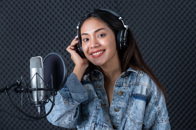 Feliz alegre muy sonriente de retrato de joven vocalista asiática con auriculares grabando una canción frente al micrófono en un estudio profesional