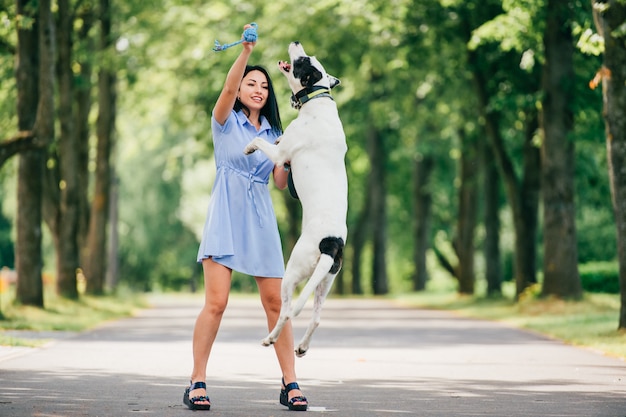 Feliz alegre niña morena sonriente en vestido azul de verano jugando con gran perro de caza en el parque