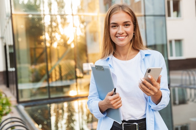 feliz alegre otimista jovem negócios com prancheta posando ao ar livre perto do centro de negócios, usando telefone celular.