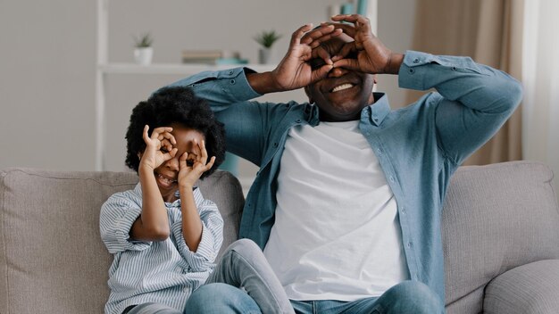 Feliz y alegre padre afroamericano maduro jugando con una linda hija sentada en la habitación en el sofá