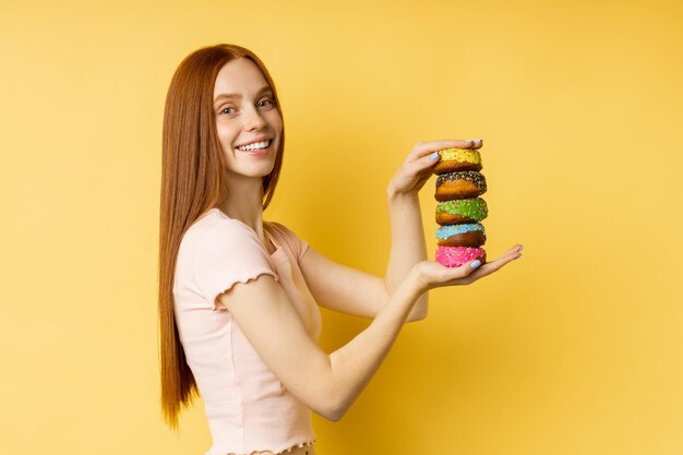 Feliz alegre pelirroja chica golosa sosteniendo un montón de coloridos donuts glaseados, sonriendo ampliamente mirando a la cámara contra la pared amarilla del estudio. Comida chatarra, pastelería, concepto de panadería.