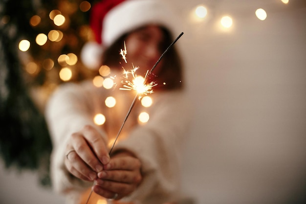 Feliz ambiente de fiesta de Nochevieja Bengala ardiendo en la mano de una chica elegante con sombrero de santa en el fondo de la luz moderna del árbol de Navidad en una habitación oscura Mujer con fuegos artificiales Felices fiestas