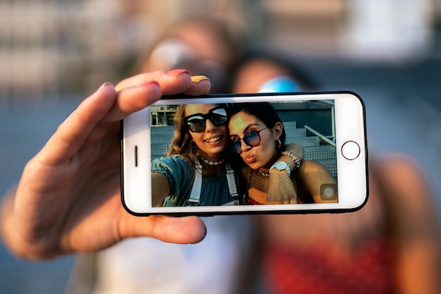 Feliz amigas jóvenes en gafas de sol sentado y tomando un selfie en el teléfono móvil de la calle de la ciudad