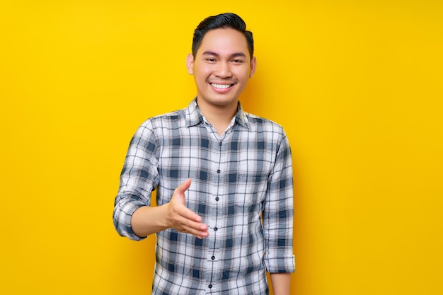 Foto feliz amistoso joven apuesto hombre asiático vestido con ropa informal mano extendida para saludar aislado sobre fondo amarillo concepto de estilo de vida de la gente