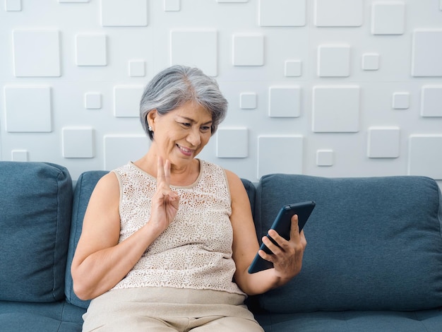 Feliz anciana asiática sentada en el sofá haciendo el signo de la victoria a mano en la pantalla de la tableta digital en la mano en la habitación blanca Las mujeres mayores se reúnen con la familia por videollamada en casa Personas mayores con tecnología