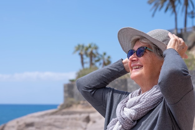 Feliz anciana caucásica con sombrero relajándose frente al mar disfrutando del verano y el horizonte de vacaciones sobre el agua