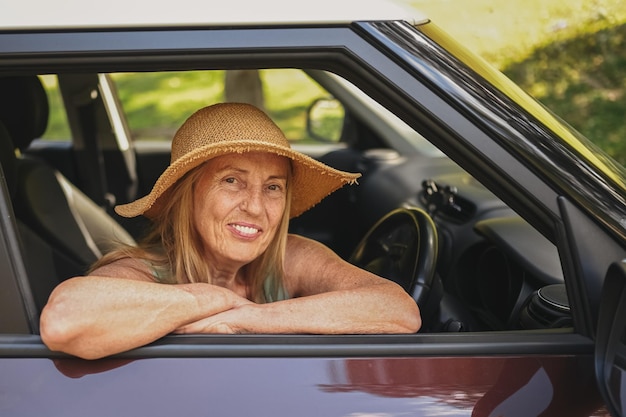 Feliz anciana conductora con sombrero de paja conduciendo sentada en un coche nuevo sonriendo mirando a la cámara disfrutando