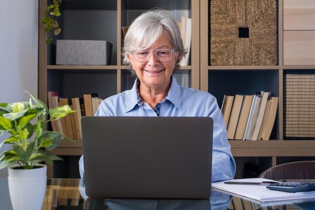 Feliz anciana empresaria caucásica sonriendo trabajando en línea viendo podcast de seminario web en una computadora portátil y aprendiendo cursos de educación llamadas en conferencia hacer notas sentarse en el escritorio elearning conceptxA