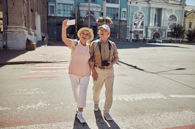 Feliz anciana fotografiándose a sí misma y a su esposo con un teléfono inteligente en el paso de peatones