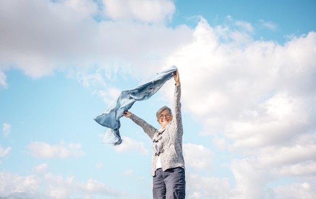 Feliz anciana madura al aire libre agitando una bufanda azul en el viento Sonriente anciana caucásica con el pelo corto con gafas disfrutando de la libertad y las vacaciones