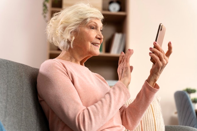 Feliz anciana madura usando la aplicación de teléfono móvil para videollamadas agitando la mano sintiéndose emocionada mientras se sienta en el sofá en casa Foto de stock