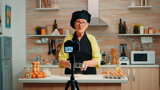 Feliz anciana panadera filmando cocinando vlog en la cocina de casa. Influencer chef blogger jubilado que usa la tecnología de Internet para comunicarse, grabar blogs en las redes sociales con equipos digitales