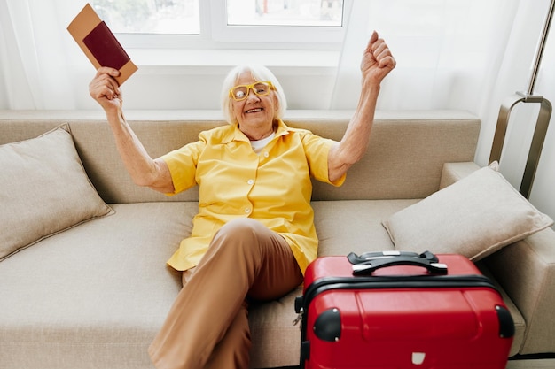 Feliz anciana con pasaporte y boleto de viaje empacó una maleta roja de vacaciones y atención médica Sonriente anciana alegremente sentada en el sofá antes del viaje levantó las manos con alegría