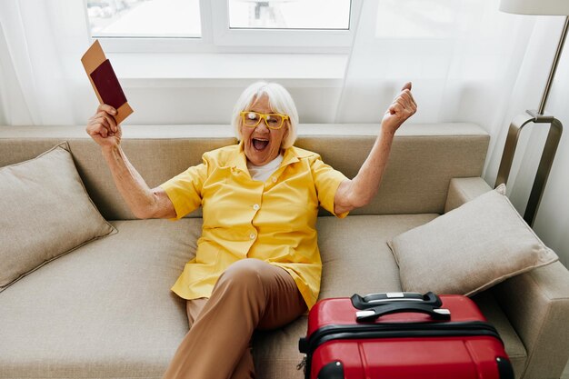 Feliz anciana con pasaporte y boleto de viaje empacó una maleta roja de vacaciones y atención médica Sonriente anciana alegremente sentada en el sofá antes del viaje levantó las manos con alegría