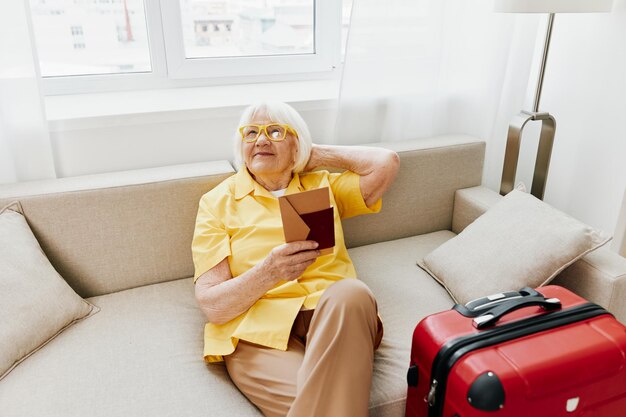 Feliz anciana con pasaporte y boleto de viaje empacó una maleta roja de vacaciones y atención médica Sonriente anciana alegremente sentada en el sofá antes del viaje levantó las manos con alegría