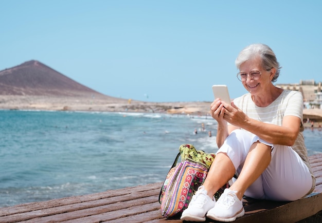 Feliz anciana sentada cerca de la playa mientras usa el teléfono móvil anciana hermosa dama disfrutando de vacaciones en el mar en un día soleado y ventoso