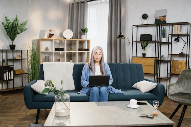 Feliz anciana sonriente sentada en un sofá azul y estudiando con una laptop en casa